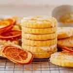 A stack of round, glazed orange shortbread cookies sits on a cooling rack, surrounded by dried orange slices and orange zest.