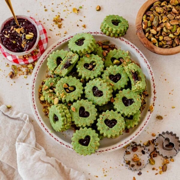 pistachio linzer cookies stacked on a Christmas plate on a counter with crushed pistachios