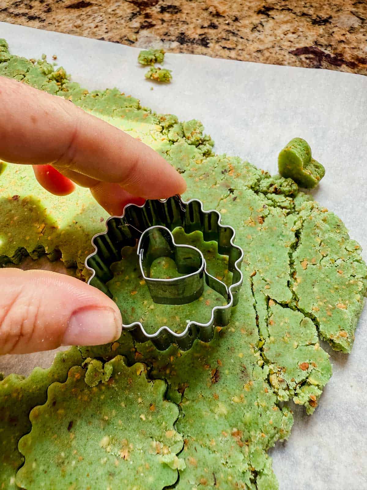 hand pressing into rolled out pistachio linzer cookie dough with a heart shaped cookie cutter