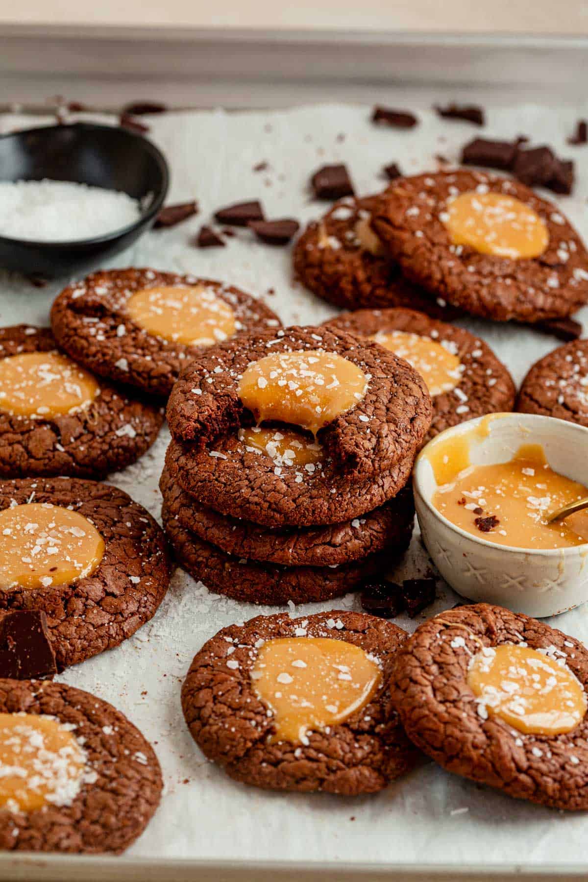 one salted caramel brownie cookie with a bite taken out of it stacked on top of a pile of salted brownie cookies laying on the counter next to a bowl of melted salted caramel