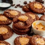 one salted caramel brownie cookie with a bite taken out of it stacked on top of a pile of salted brownie cookies laying on the counter next to a bowl of melted salted caramel