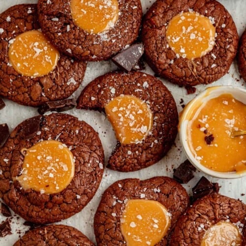 salted caramel brownie cookies sprinkled with sea salt laid out on a counter with a bite taken out of the middle one and a bowl of melted caramel