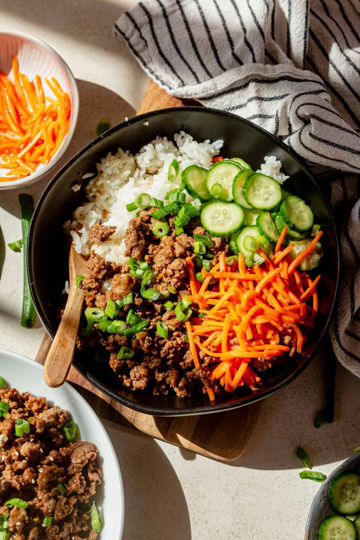 a black bowl with sticky rice, korean ground beef, cucumbers, and carrots.