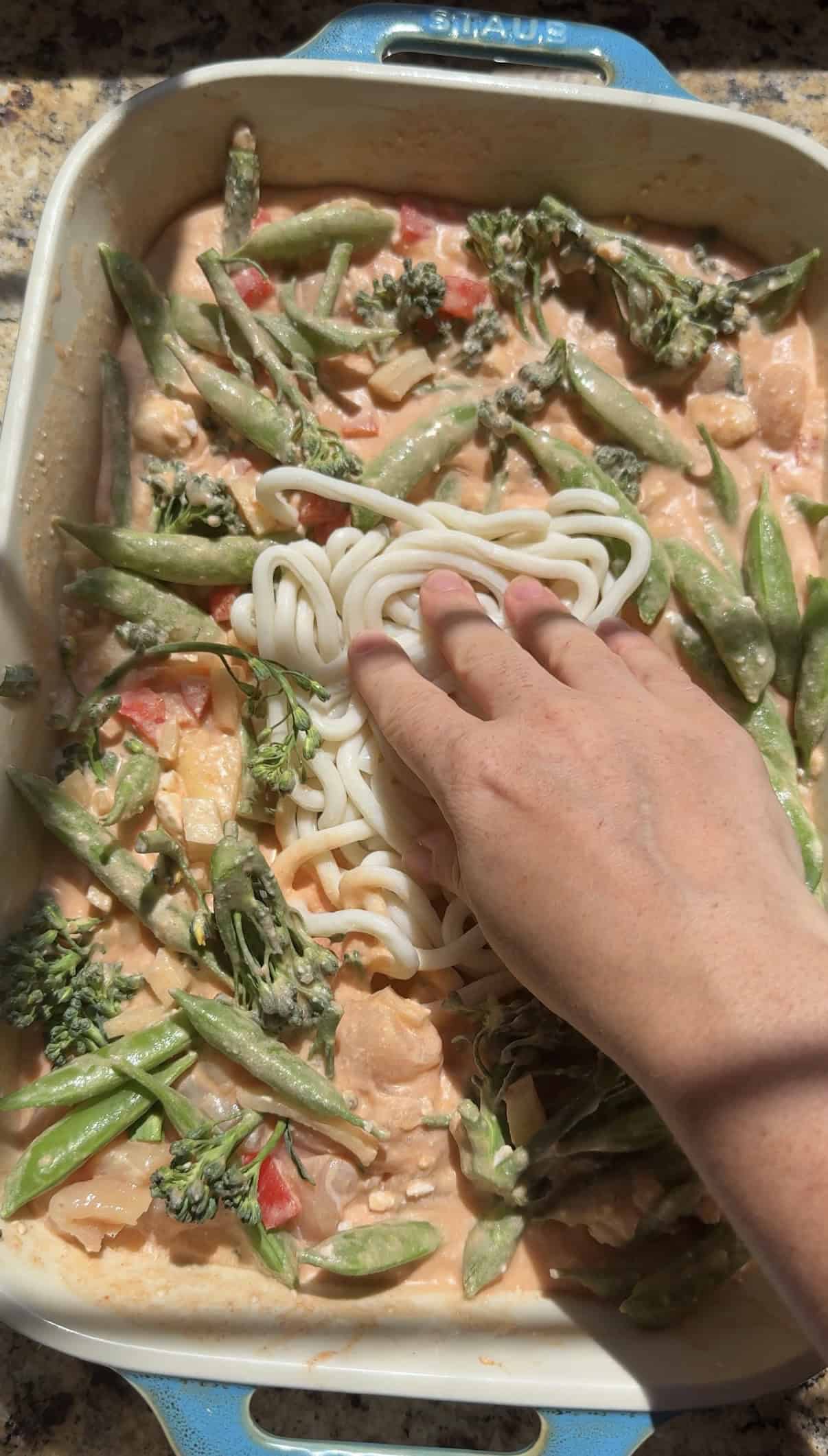 pressing udon noodles into a casserole dish of coconut curry chicken and vegetables.