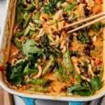 Square crop of chicken curry udon noodles being lifted with chopsticks from the baking dish, showing the glossy red curry sauce, snap peas, charred broccolini, and cilantro.