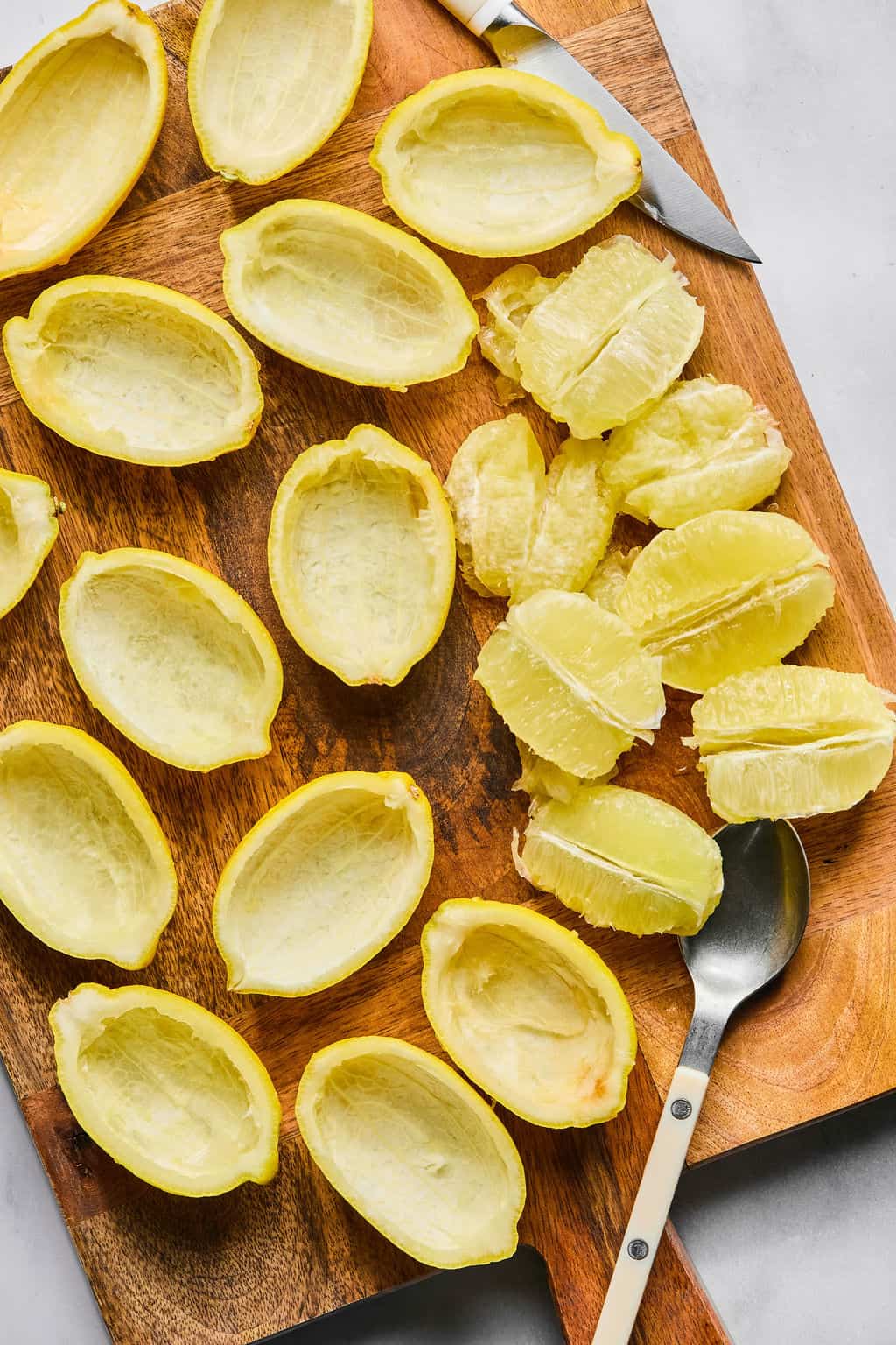 Empty yellow citrus peels and peeled citrus segments are arranged on a wooden cutting board. A small paring knife and a spoon with a white handle are placed nearby.