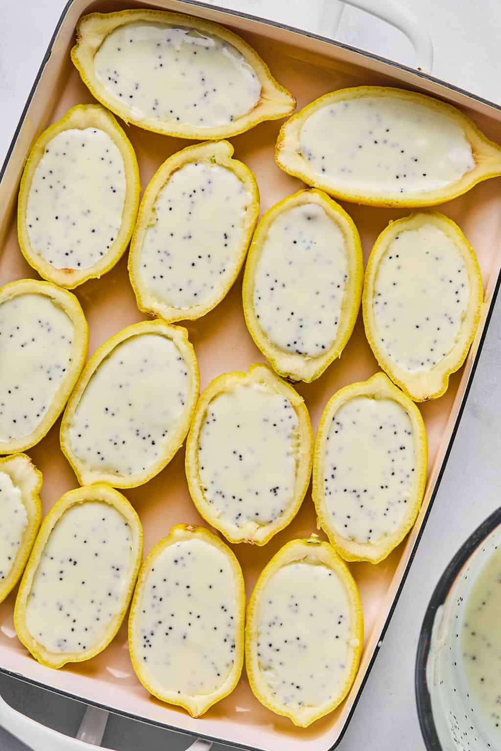 A baking dish filled with lemon rind halves, each filled with a creamy lemon posset containing poppy seeds. The pastries are neatly arranged in rows, ready to be chilled.