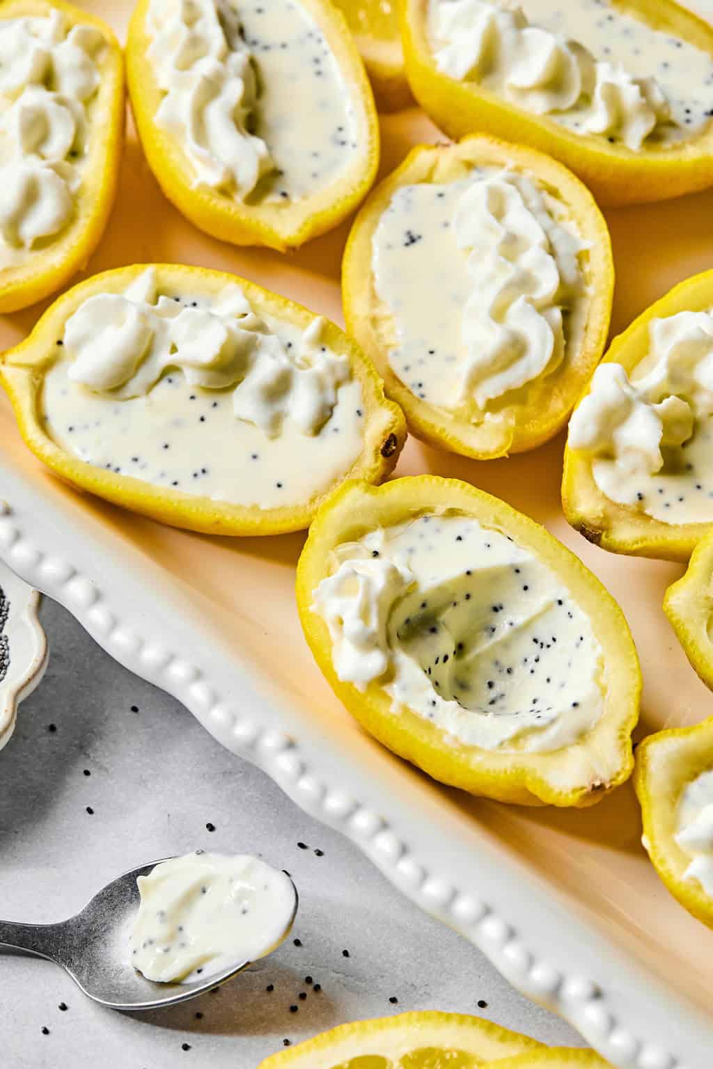 Halved lemons filled with creamy lemon poppyseed posset, topped with whipped cream and sprinkled with poppy seeds, are arranged on a white tray. A spoon with some of the dessert rests nearby.