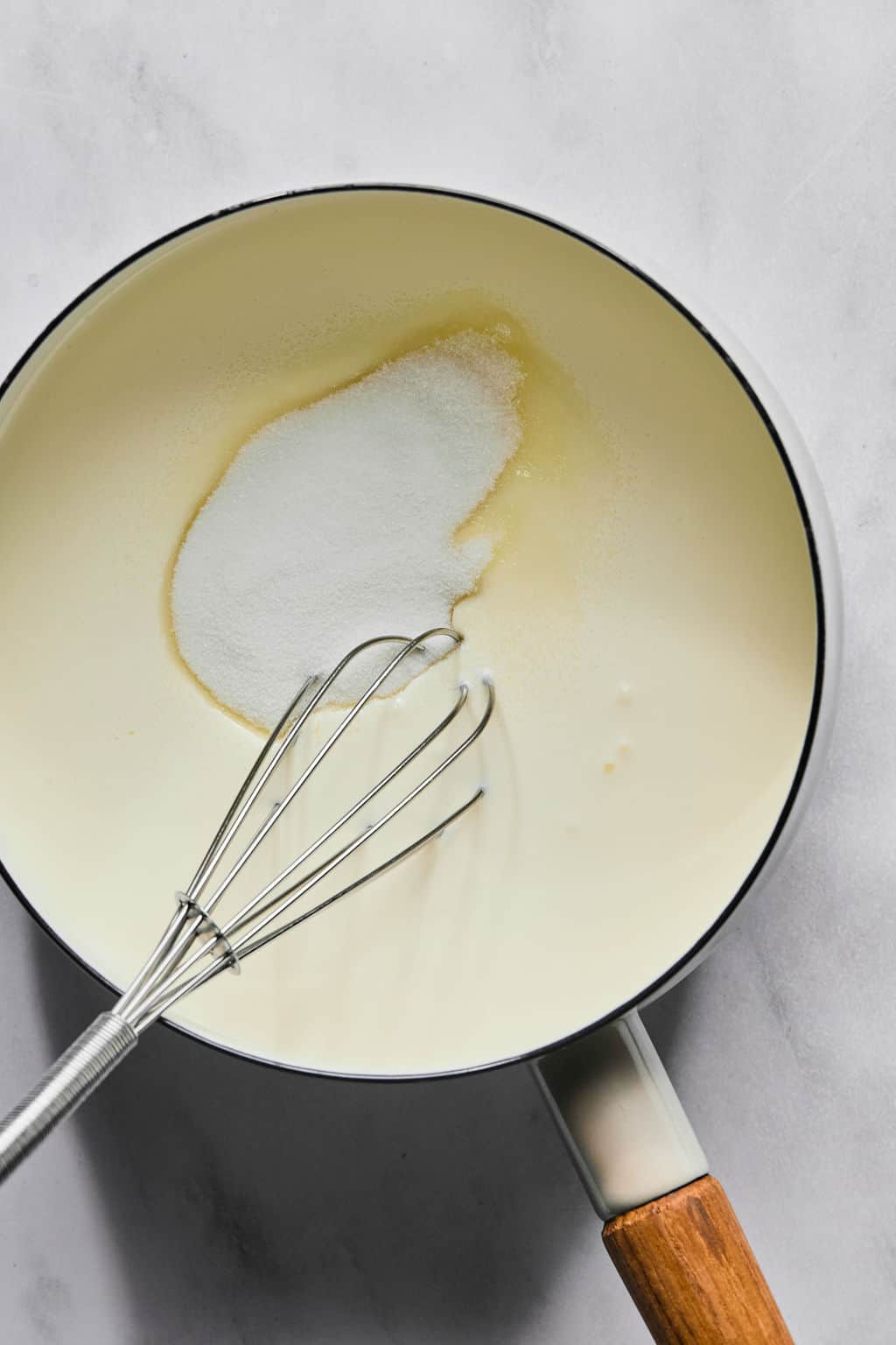 A saucepan with a wooden handle contains cream and a mound of sugar, being mixed with a metal whisk, on a light-colored surface.