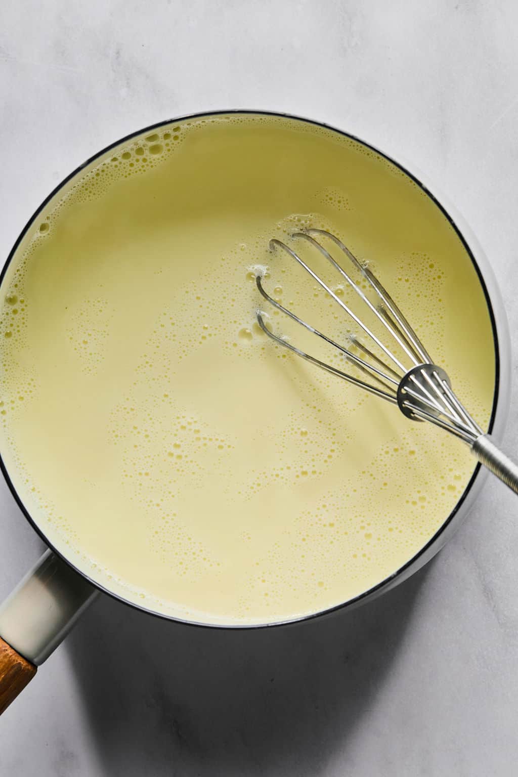 A saucepan filled with a creamy, pale yellow custard mixture being stirred by a metal whisk, placed on a light gray surface.