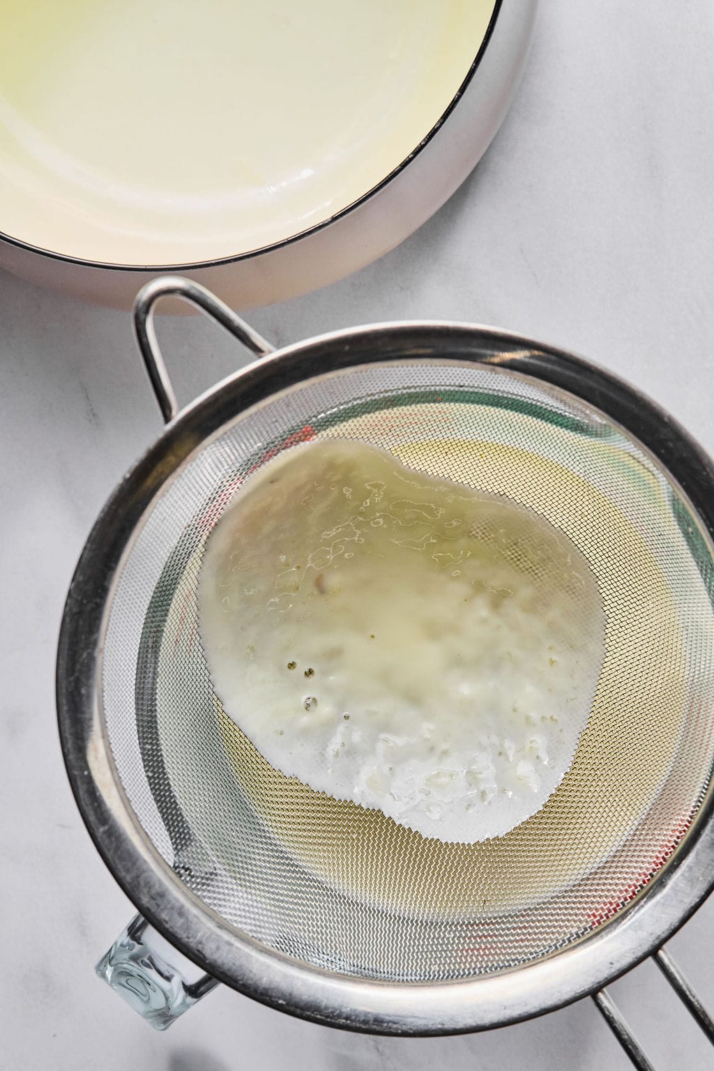 A metal strainer holding foamy liquid is placed over a glass bowl, with a white saucepan partially visible in the background on a white surface.