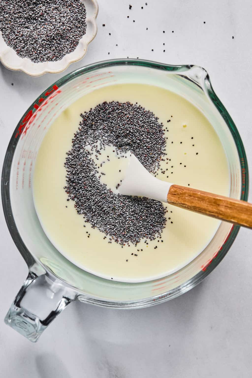 A glass measuring cup filled with lemon posset mixture, with poppy seeds being stirred in using a white spatula with a wooden handle. A small dish of extra poppy seeds is nearby on a white surface.