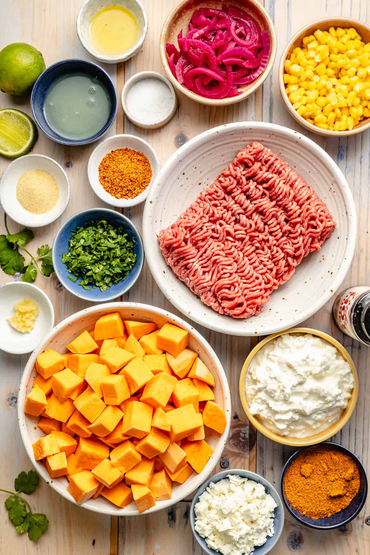 Overhead view of various ingredients in bowls on a wooden surface, including ground beef, cubed sweet potatoes, corn, pickled onions, cottage cheese, lime halves, cilantro, feta, and assorted spices.