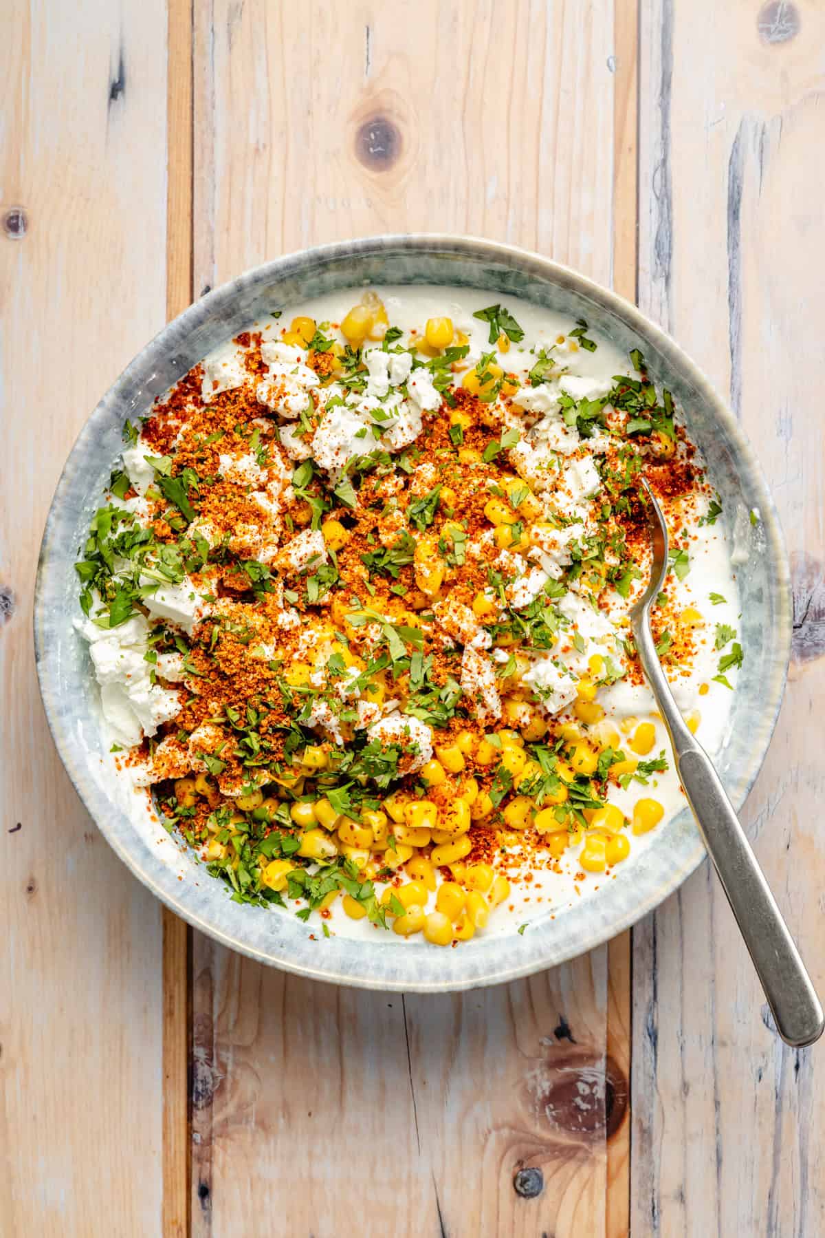 A bowl filled with corn, crumbled cheese, chopped cilantro, and red chili powder on a creamy base, with a spoon placed inside. The bowl sits on a light wooden surface.
