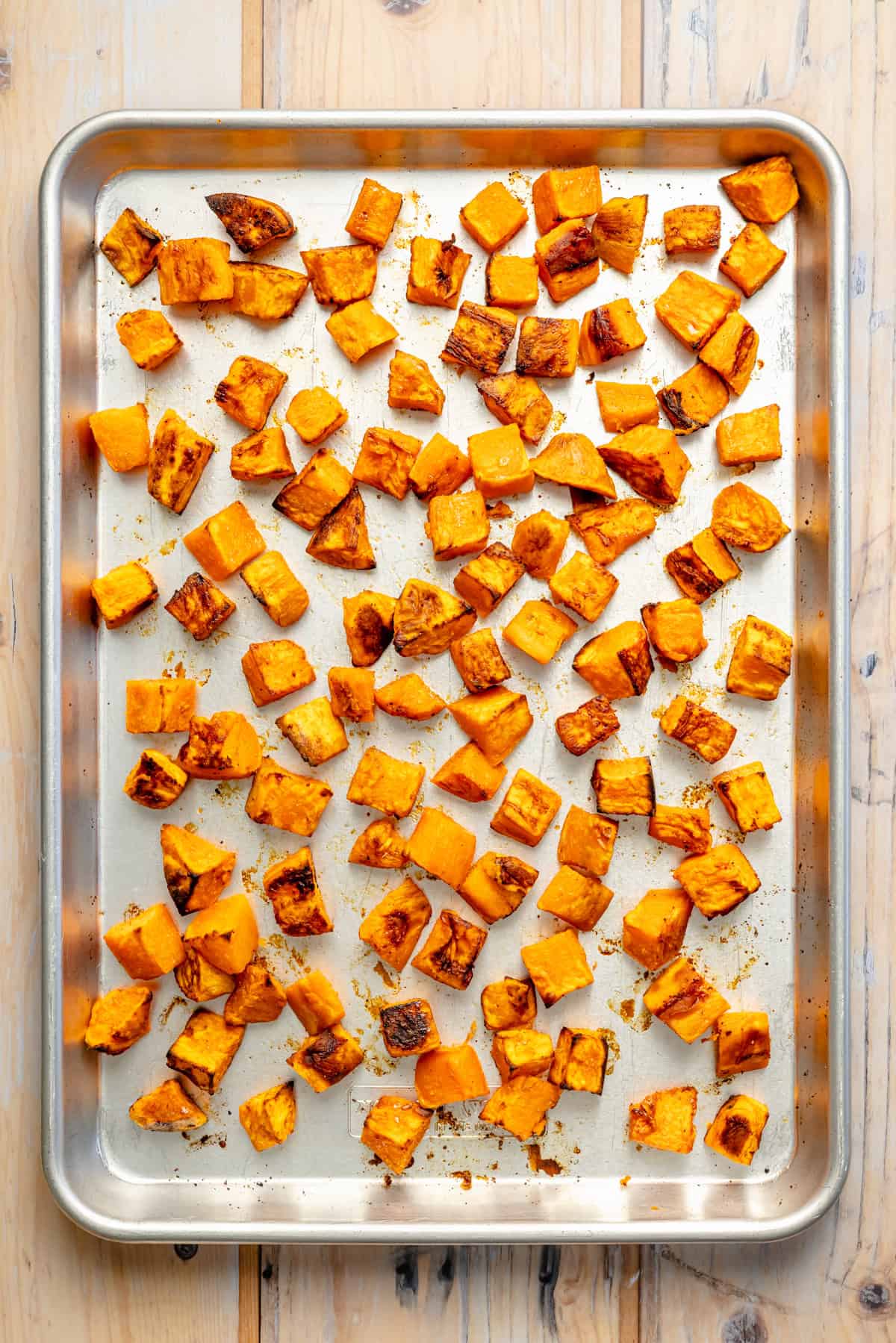 Roasted sweet potato cubes spread evenly on a metal baking sheet, showing golden brown edges. The tray rests on a light wood surface.