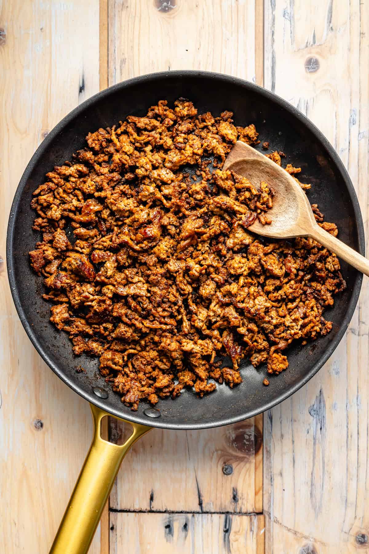 A skillet filled with cooked, seasoned ground meat and a wooden spoon resting on the side, placed on a light wooden surface.
