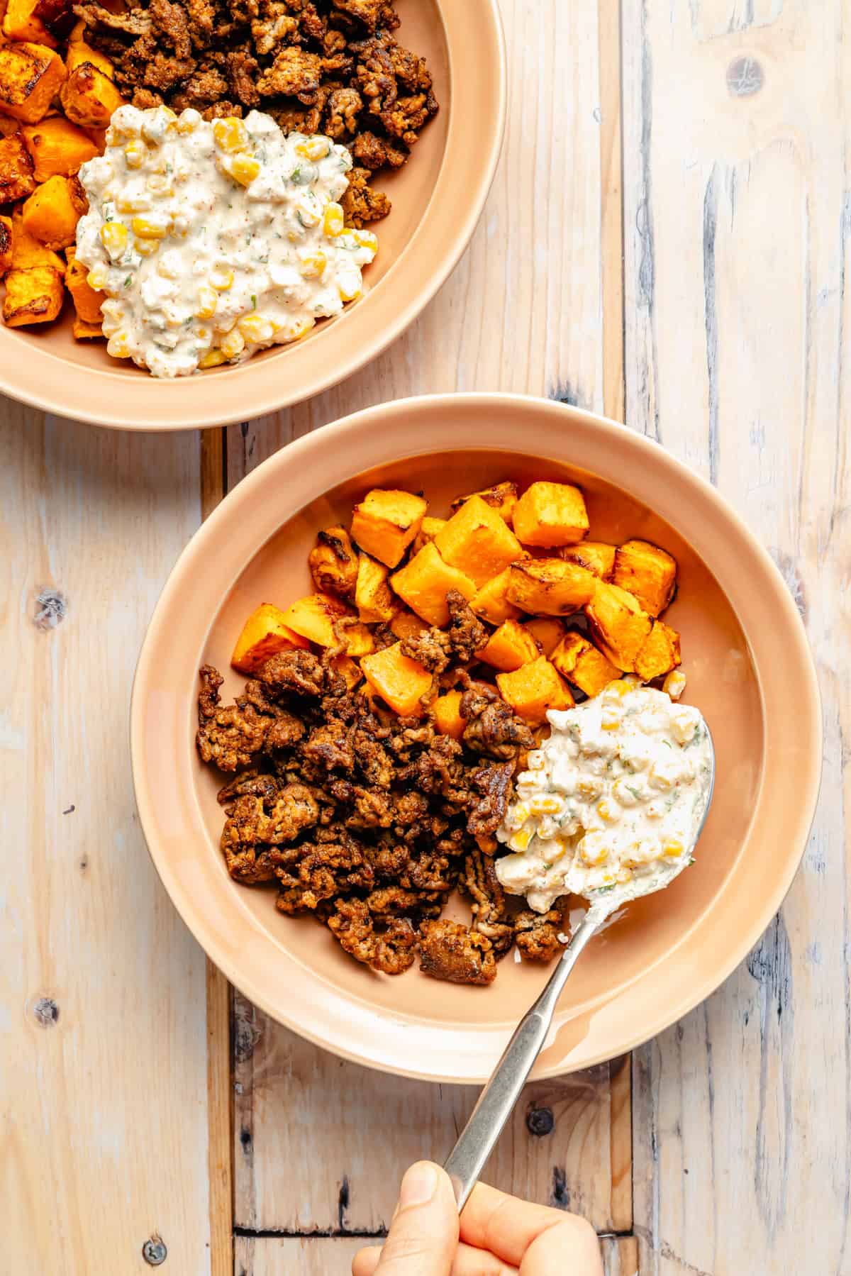 Two beige bowls with roasted sweet potatoes and browned ground meat, topped with a creamy corn mixture. A hand holds a spoon, scooping the corn topping onto one bowl on a light wooden table.