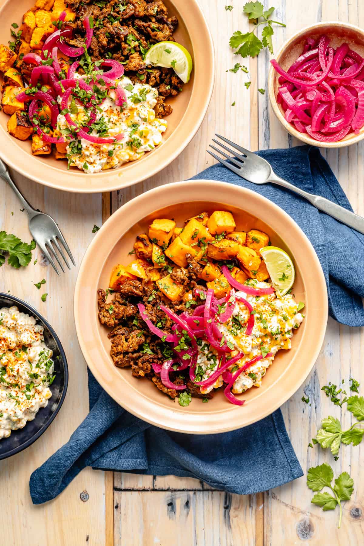 Two bowls filled with ground meat, roasted sweet potatoes, corn, pickled red onions, cilantro, and lime wedges, set on a wood table with a blue napkin, forks, and a small bowl of extra pickled onions nearby.