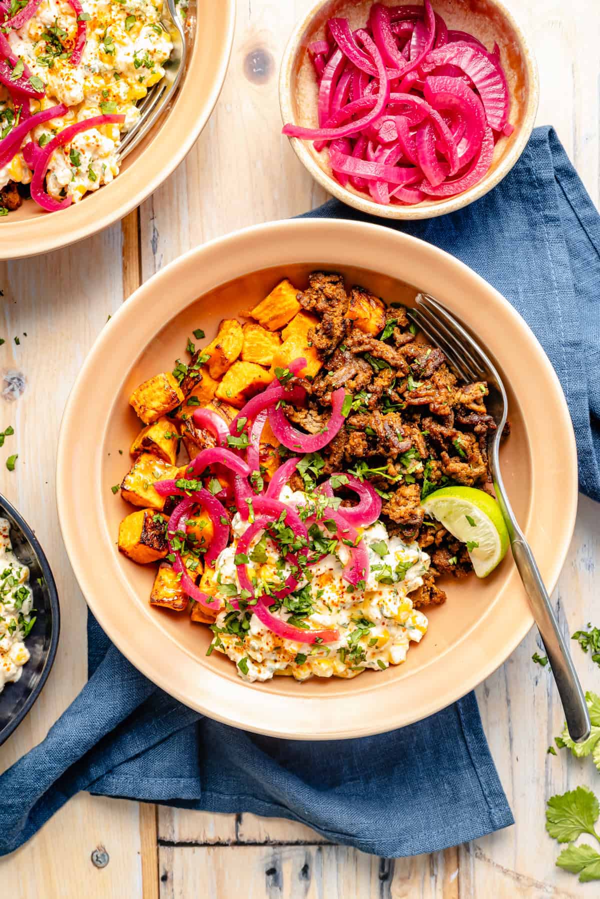 A bowl with seasoned ground meat, roasted sweet potatoes, Mexican street corn salad, and pickled red onions, garnished with cilantro and a lime wedge. A fork rests on the side; other bowls and onions are nearby on a wooden table.
