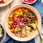A bowl with seasoned ground meat, roasted sweet potatoes, pickled red onions, cilantro, a lime wedge, and a creamy corn salad, served on a tan plate with a fork, on a blue napkin.