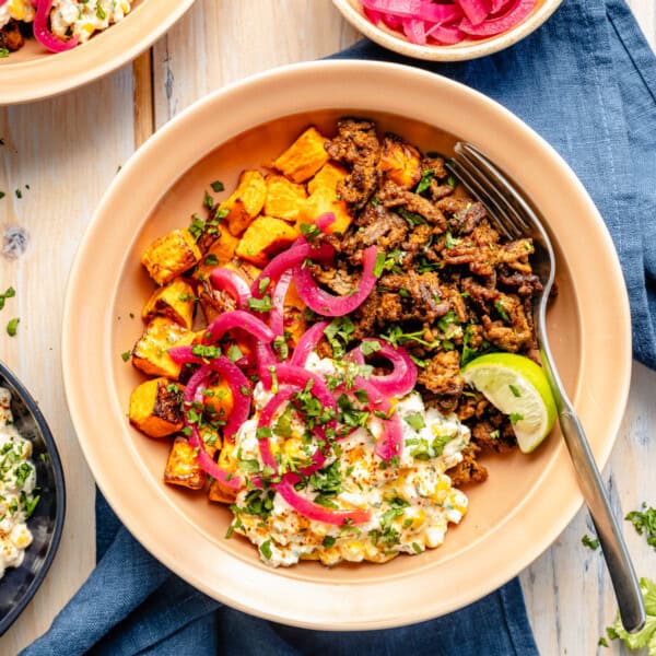 A bowl with seasoned ground meat, roasted sweet potatoes, pickled red onions, cilantro, a lime wedge, and a creamy corn salad, served on a tan plate with a fork, on a blue napkin.