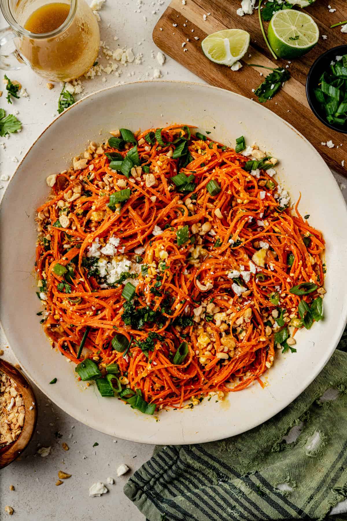 carrot ginger salad tossed in dressing in a white bowl with a cutting board, fresh limes, and a green towel surrounding it.