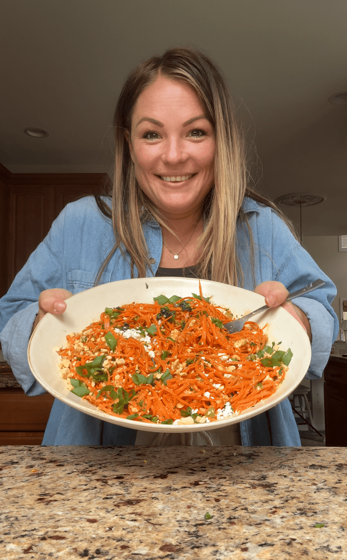 molly thompson holding a white serving bowl full of carrot ginger salad with a serving spoon resting in it.