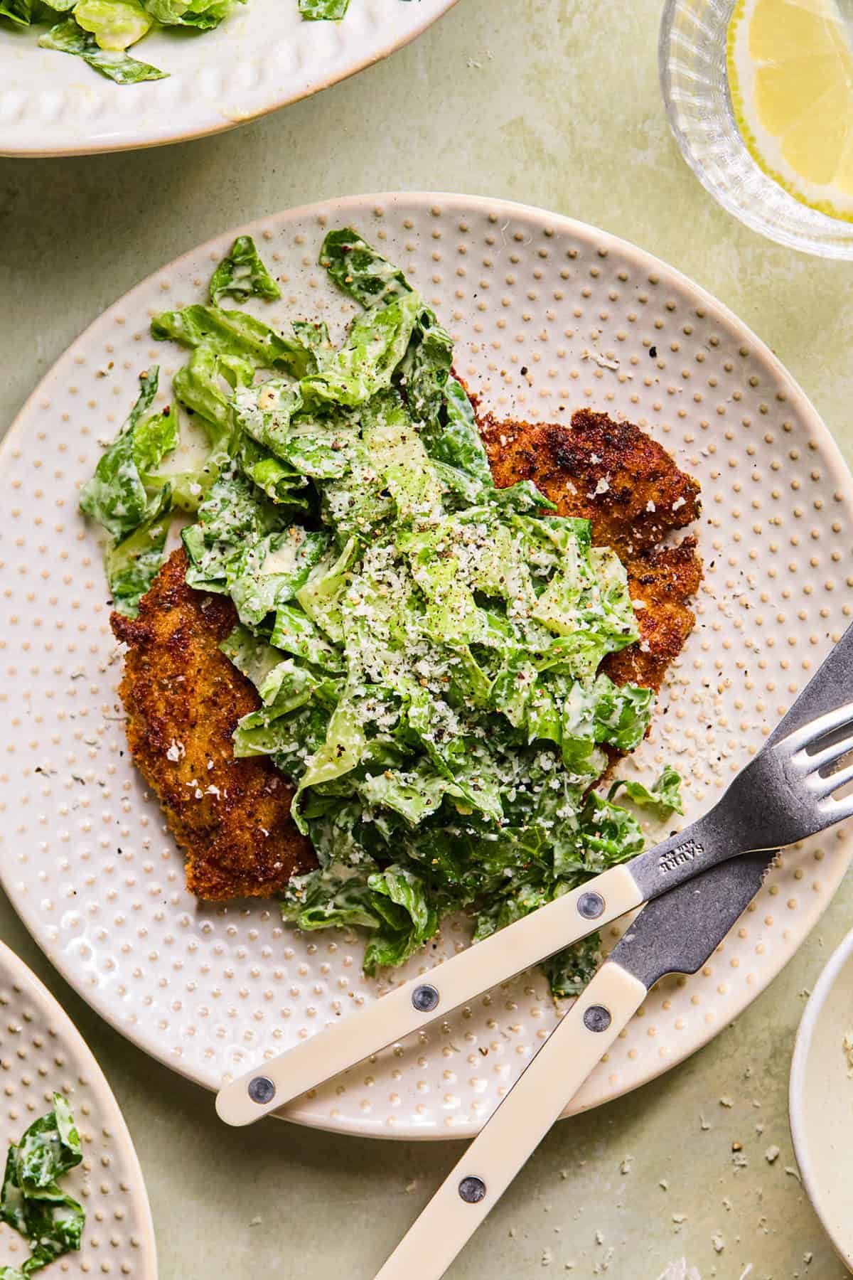 A breaded, pan-fried chicken cutlet topped with chopped romaine lettuce and grated cheese is served on a ceramic plate with a fork and knife beside it. A slice of lemon and more salad are visible nearby.