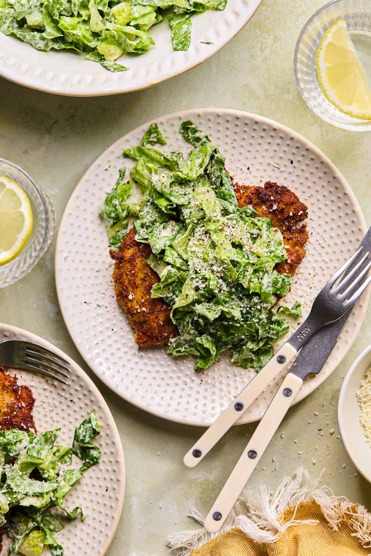 A plate with breaded crispy chicken cutlets topped with creamy caesar salad, beside a fork and knife. Surrounding plates and glasses with lemon slices are also visible on a light green surface.