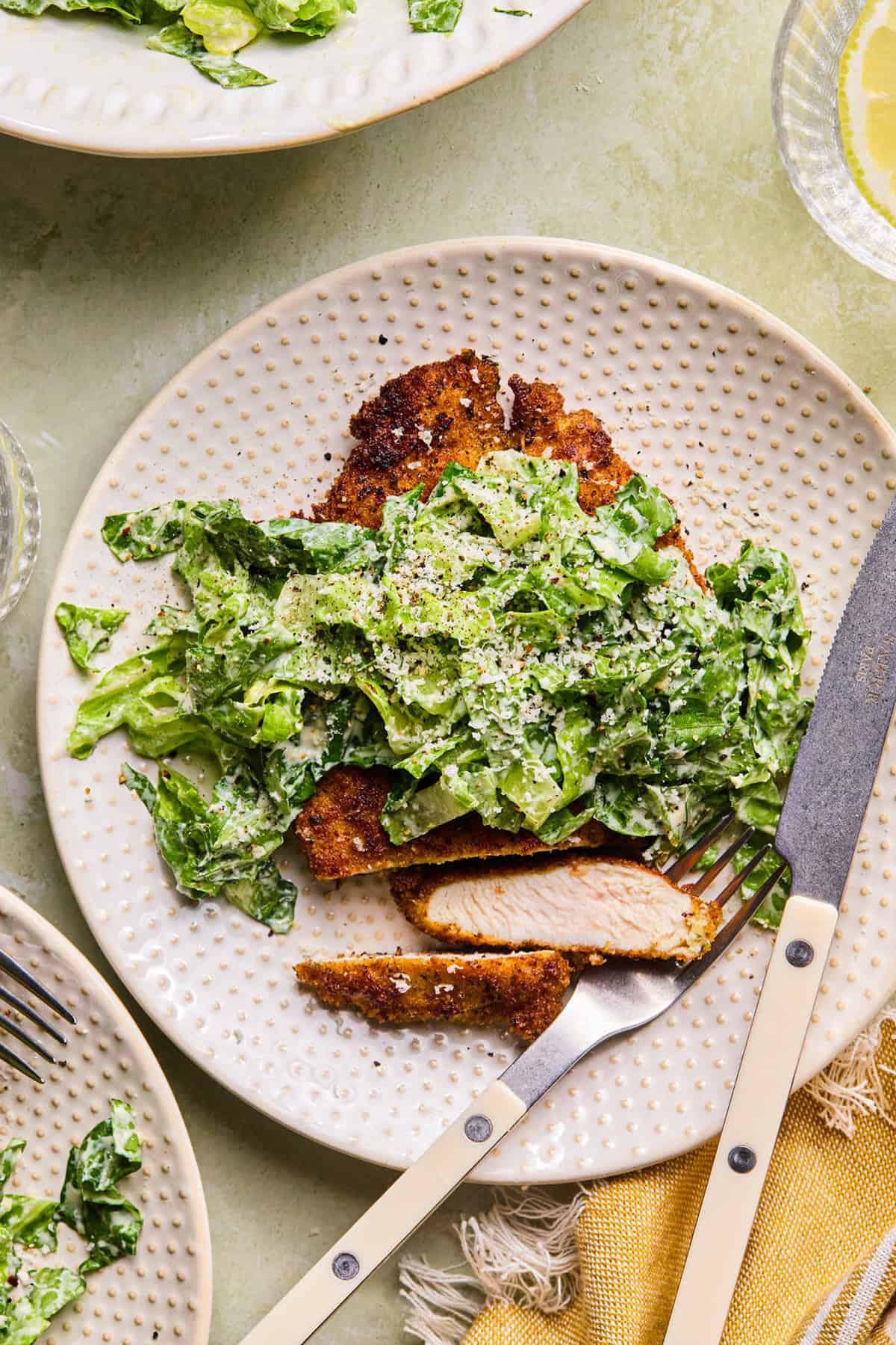 A plate with breaded, sliced chicken topped with chopped romaine lettuce, grated cheese, and black pepper. A fork and knife rest on the plate, and part of a yellow napkin is visible beside it.