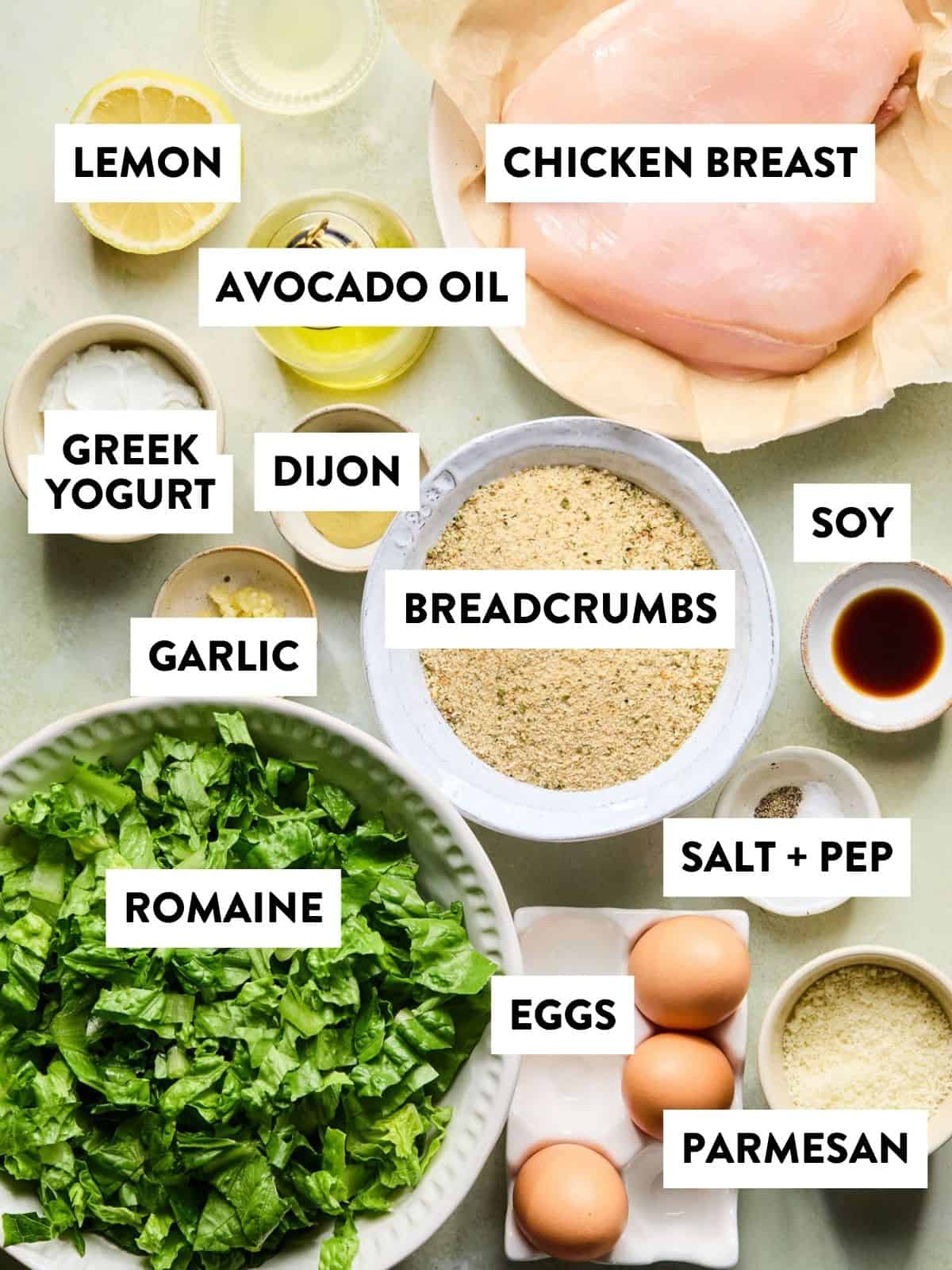 Top-down view of labeled ingredients for a crispy chicken Caesar recipe, including chicken breast, lemon, avocado oil, Greek yogurt, Dijon, breadcrumbs, soy sauce, garlic, romaine lettuce, salt and pepper, eggs, and parmesan.