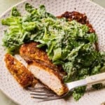 A plate with crispy breaded chicken cutlets and a fresh green caesar salad topped with grated parmesan cheese. A fork rests on the plate with a piece of chicken speared on it.