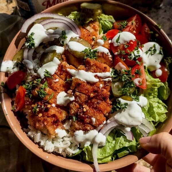 A bowl of rice topped with crispy fried chicken, shredded lettuce, sliced red onions, cherry tomatoes, pickles, fresh herbs, and drizzled with creamy white sauce.