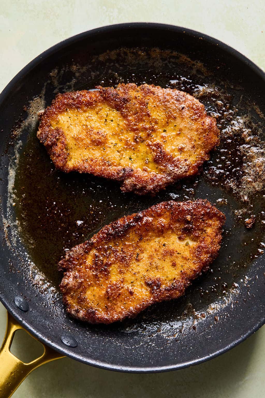 Two browned and crispy breaded cutlets are frying in a black skillet with oil, showing a golden crust and sizzling edges.