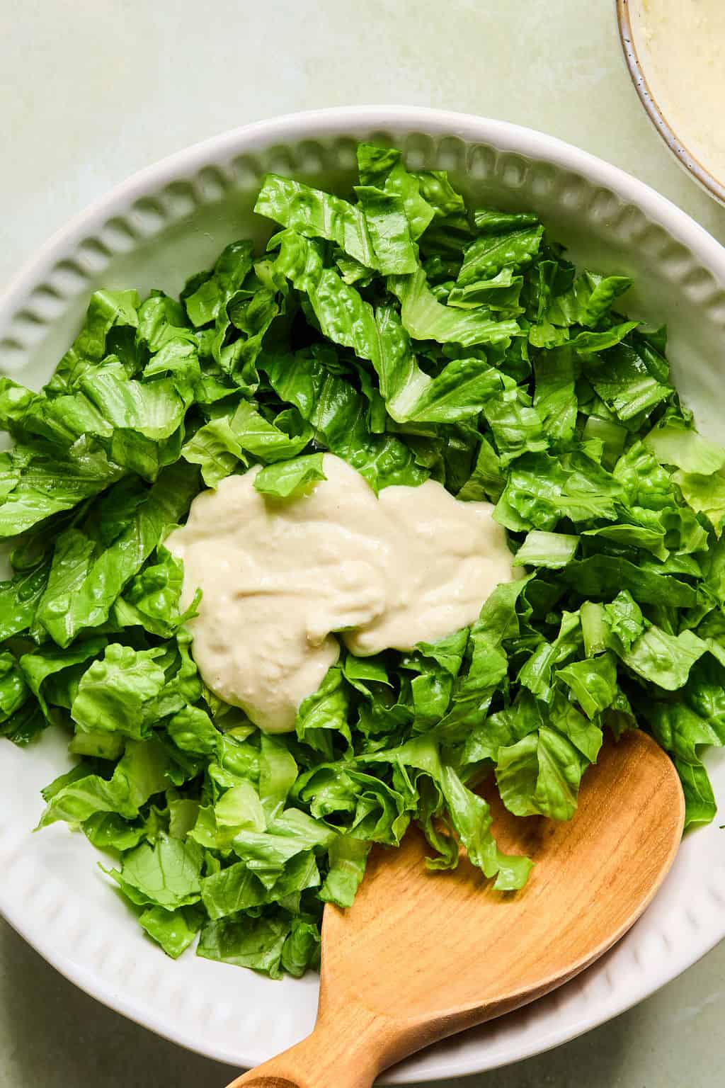 A white bowl filled with chopped romaine lettuce, topped with creamy caesar dressing, with a wooden spoon resting in the salad.