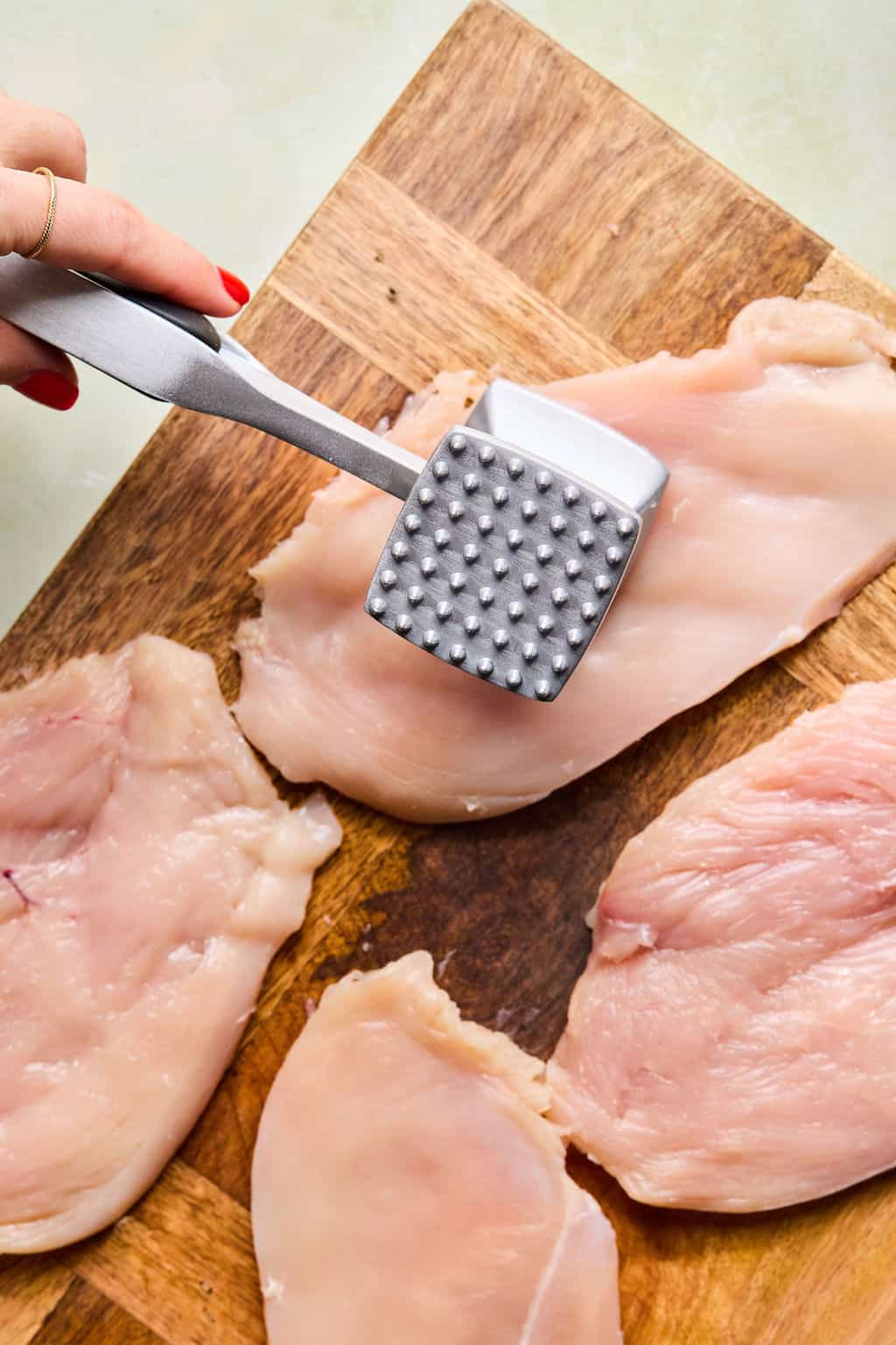 A hand uses a metal meat tenderizer to pound raw chicken breasts on a wooden cutting board. Several pieces of chicken are spread out on the board.