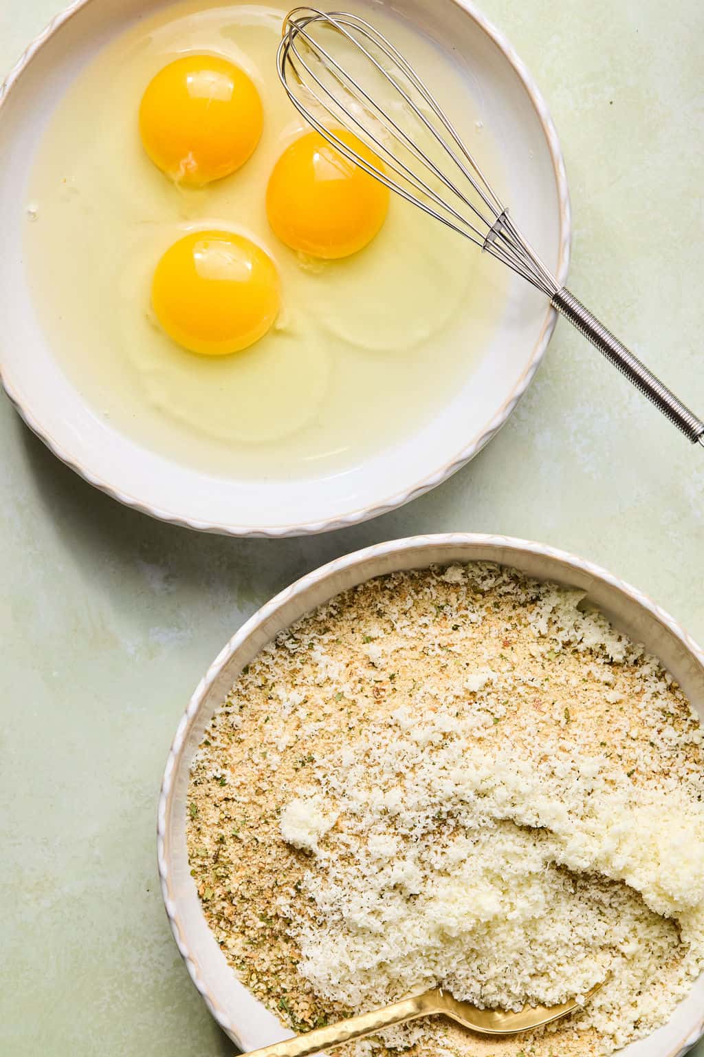 Two bowls on a light surface: one with three raw eggs and a whisk, the other with a mixture of breadcrumbs and grated cheese, with a spoon resting inside.