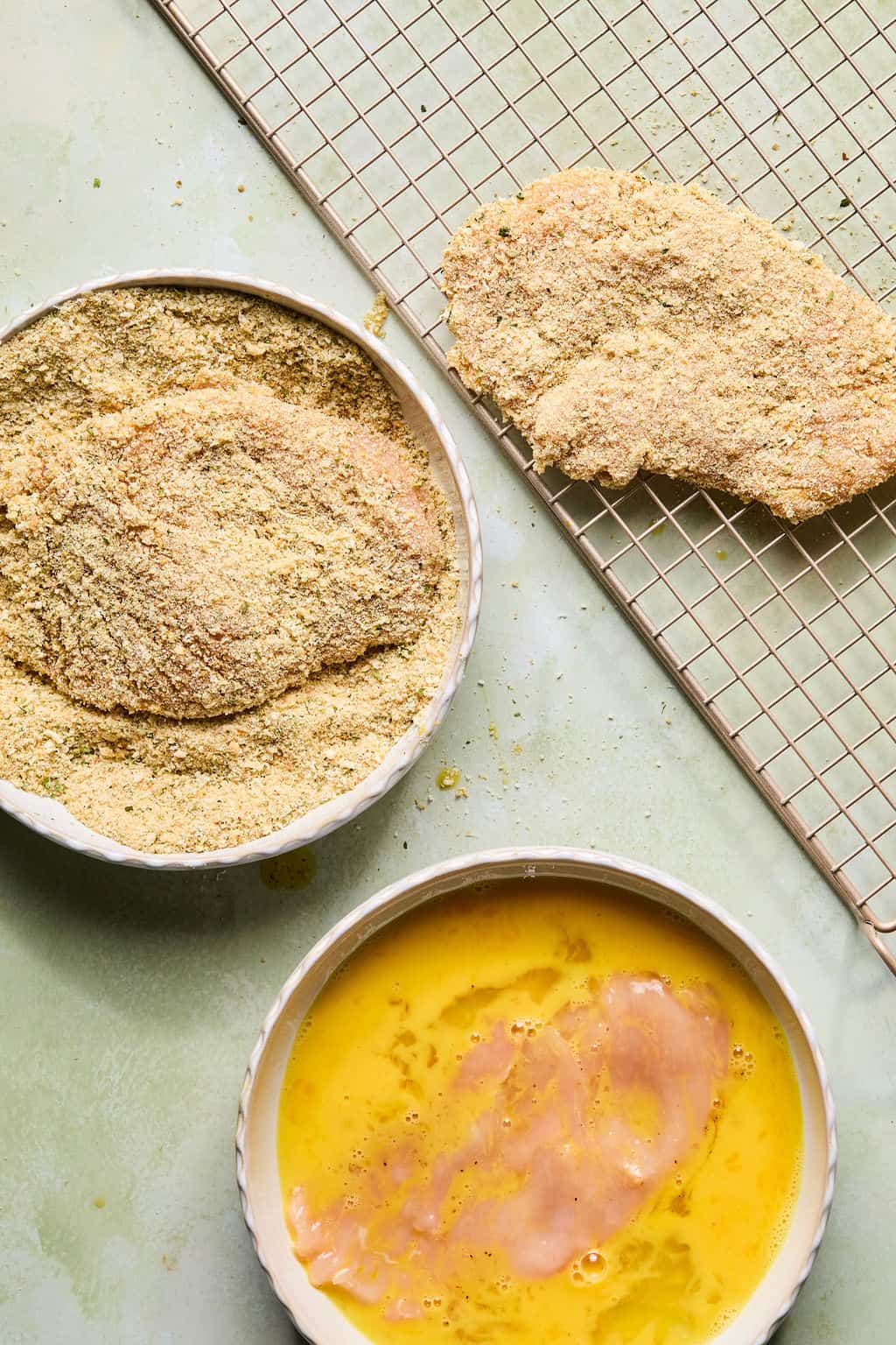 A raw chicken breast is dipped in beaten eggs, then coated in breadcrumbs. One coated piece rests on a cooling rack, while bowls with egg wash and breadcrumbs are nearby on a light green surface.