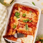 A baked cottage cheese manicotti in a white dish, topped with melted cheese and fresh basil leaves. A slice has been served, revealing layers of pasta and sauce. A knife rests on the dish, with cheese and a green towel nearby.
