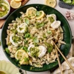 A green plate of fusilli pasta topped with dollops of cheese, pesto sauce, fresh basil leaves, and lemon slices, with gold utensils on the side. Lemon wedges, pesto, and garlic are visible around the plate.