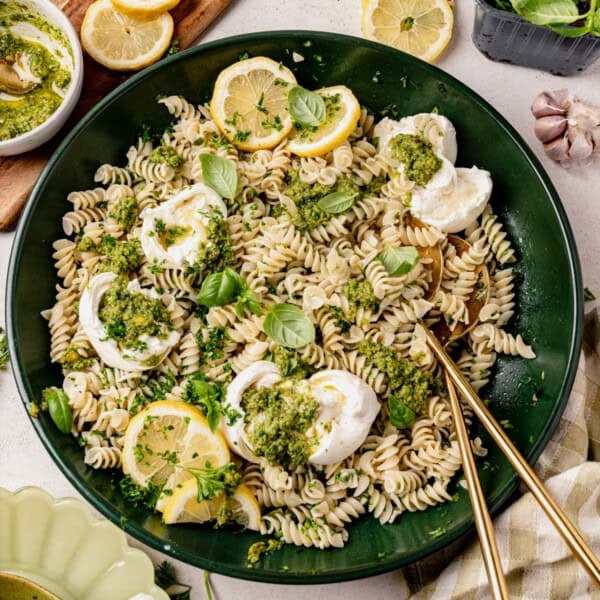 A green plate of fusilli pasta topped with dollops of cheese, pesto sauce, fresh basil leaves, and lemon slices, with gold utensils on the side. Lemon wedges, pesto, and garlic are visible around the plate.