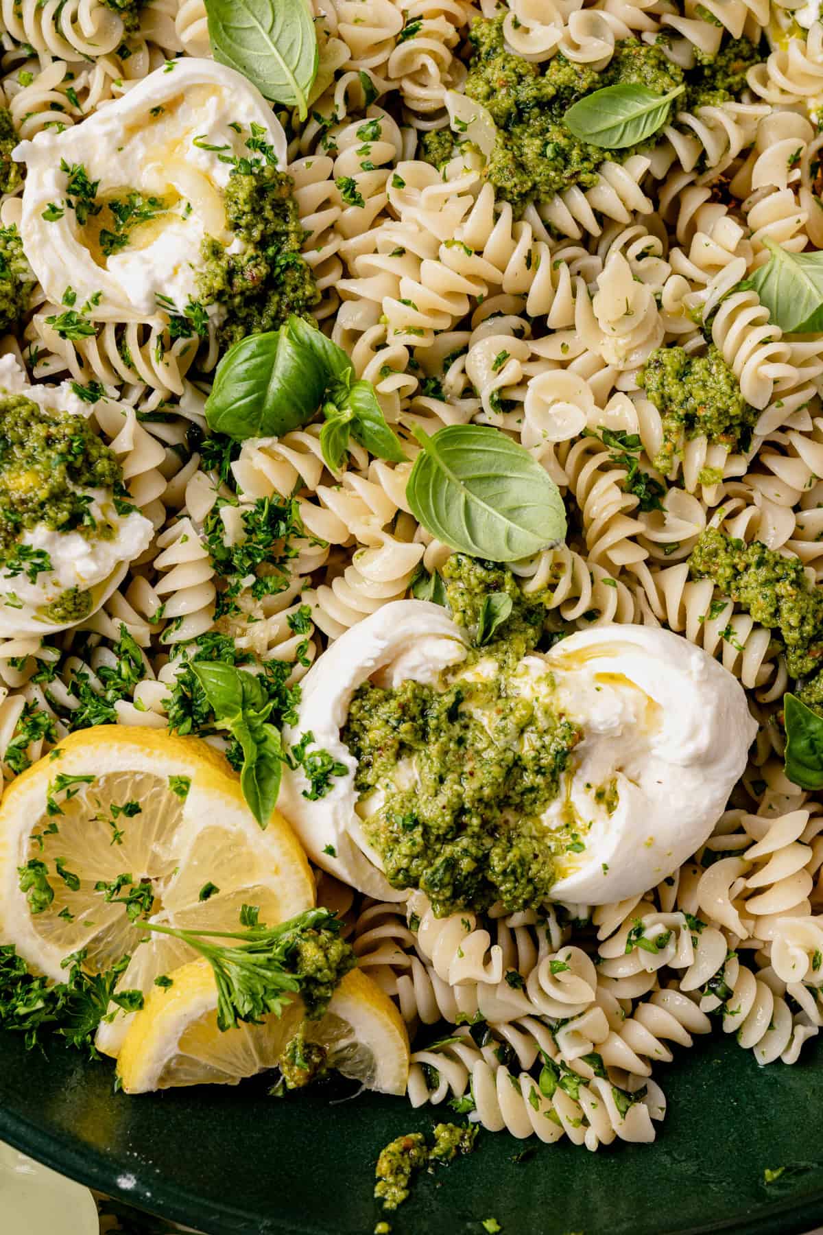 A close-up of rotini pasta topped with dollops of burrata cheese, pesto sauce, fresh basil leaves, chopped parsley, and lemon slices. The dish has a vibrant and fresh appearance.