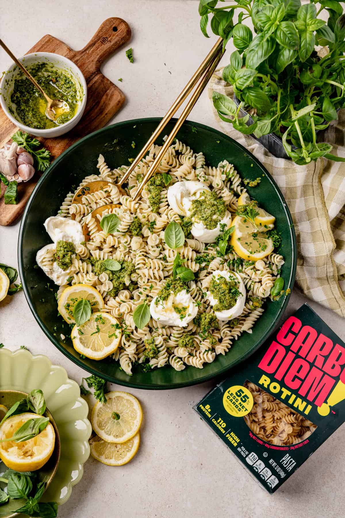 A bowl of rotini pasta with pesto, fresh mozzarella, lemon slices, and basil, surrounded by scattered ingredients, a box labeled Carbe Diem Rotini, and a small bowl of pesto with a spoon.