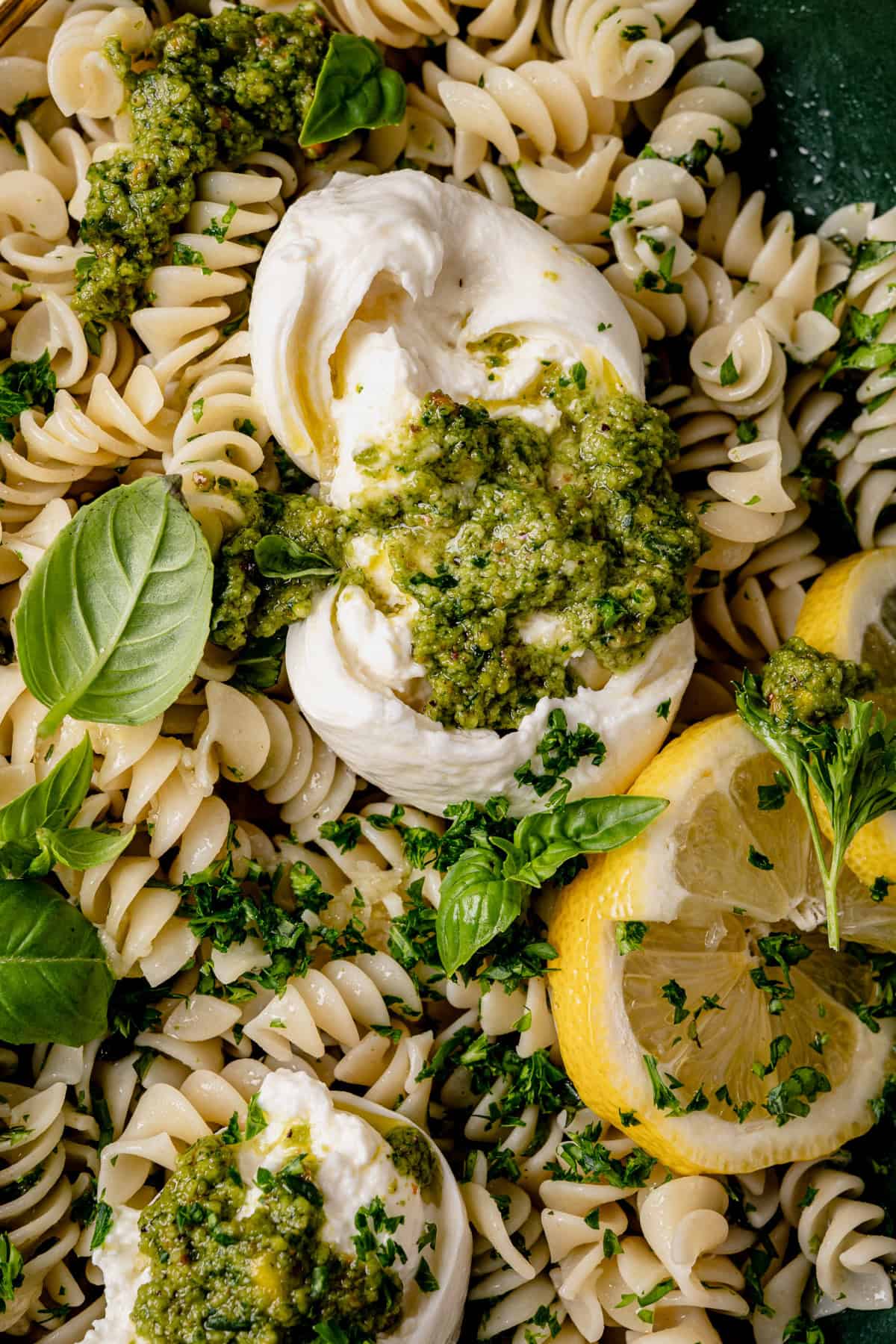 Close-up of rotini pasta topped with burrata cheese, green pesto, fresh basil leaves, chopped parsley, and lemon slices. The dish appears fresh and vibrant, showcasing a mix of green and yellow accents.