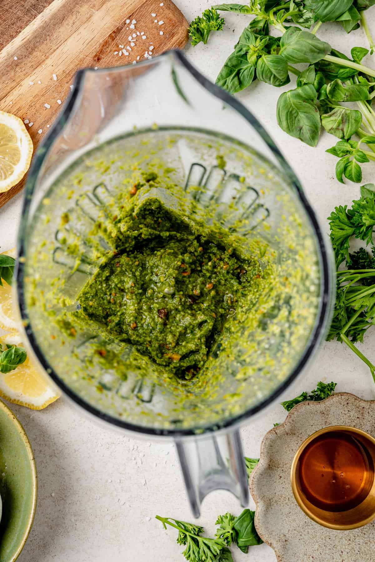 A blender filled with bright green pistachio pesto sauce, surrounded by fresh herbs, lemon slices, a wooden board, and a small bowl of oil on a white countertop.