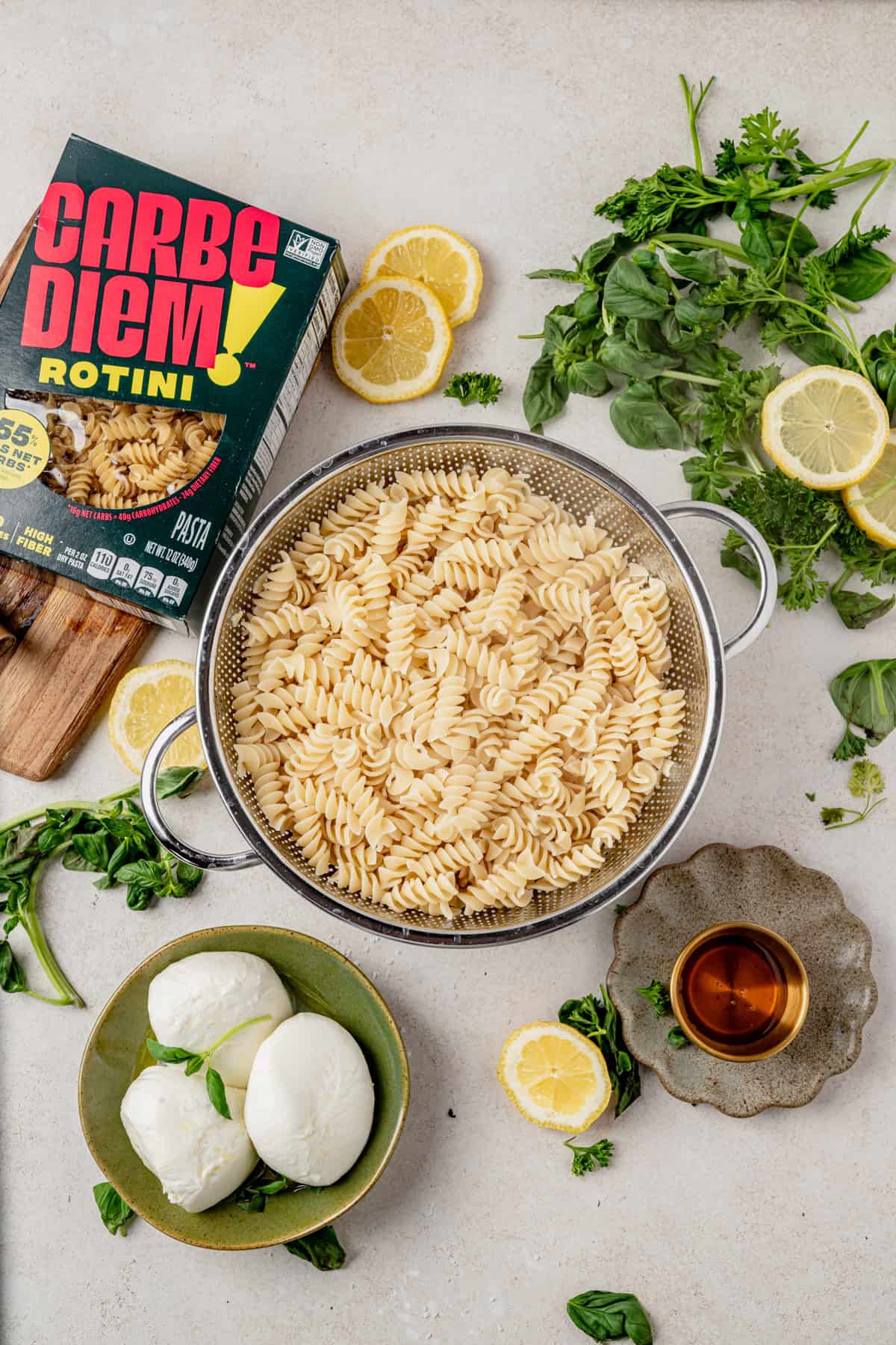 A colander filled with cooked rotini pasta sits on a countertop surrounded by a Carbe Diem Rotini box, fresh basil, mozzarella balls, lemon slices, greens, and a small bowl of olive oil.