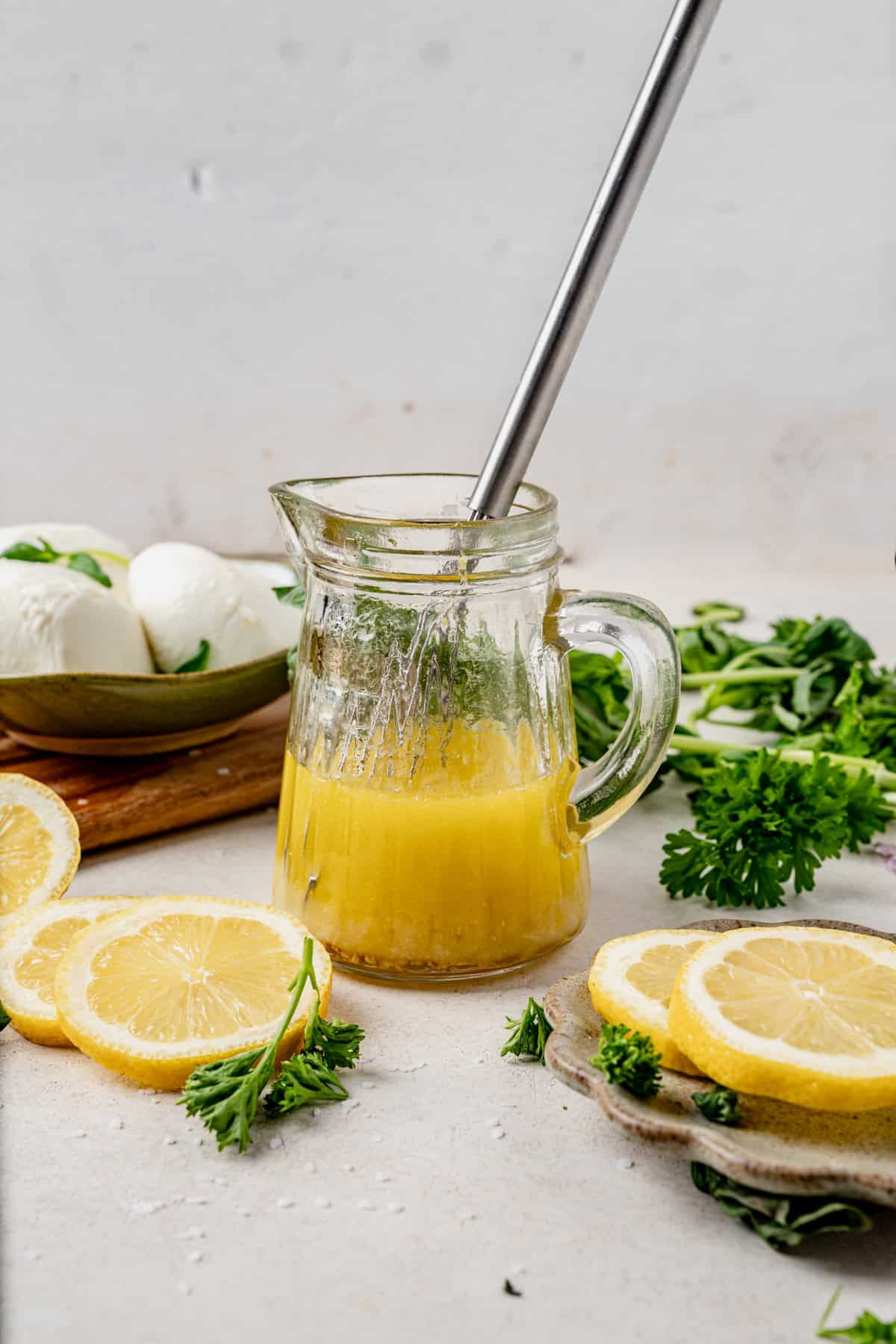 A glass pitcher with yellow dressing and a metal whisk inside, surrounded by fresh lemon slices, parsley, and a bowl of mozzarella balls on a light surface.