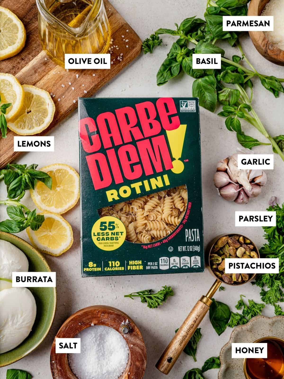A box of Carbe Diem Rotini pasta surrounded by labeled ingredients: olive oil, Parmesan, basil, lemons, garlic, parsley, pistachios, honey, burrata, and salt, all arranged on a white surface.