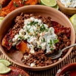 A street corn sweet potato bowl featuring seasoned ground beef over roasted sweet potato, topped with creamy corn salad and fresh herbs, served with lime wedges and extra salad in the background.