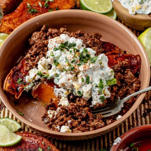 A street corn sweet potato bowl featuring seasoned ground beef over roasted sweet potato, topped with creamy corn salad and fresh herbs, served with lime wedges and extra salad in the background.
